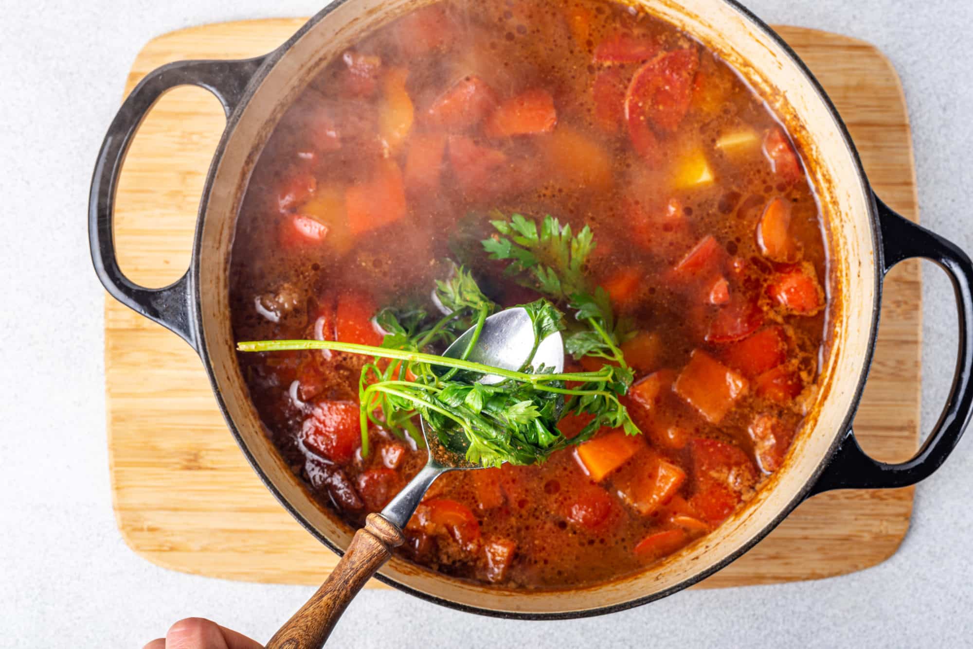 Hearty soup cooking in a large pot on a wooden board with green parsley being added in with a spoon.