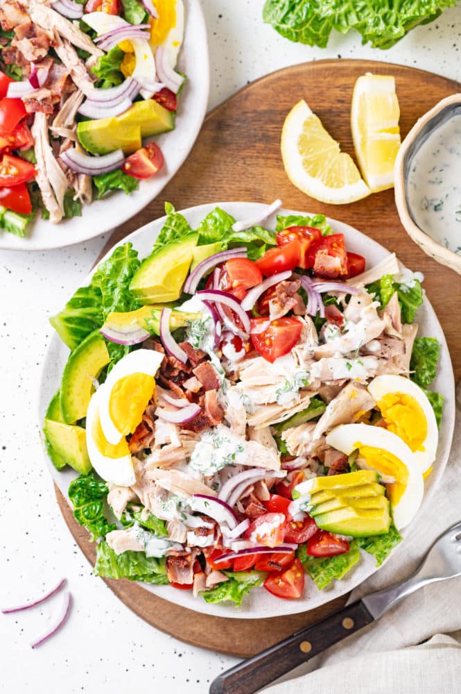 cobb-salad-chicken-tomatoes-red-onion-bacon-hard-boiled-eggs-avocado-lettuce-and-ranch-dressing-on-a-white-plate-and-wooden-board-with-a-silver-fork-and-lemon-wedges-and-bowl-of-dressing