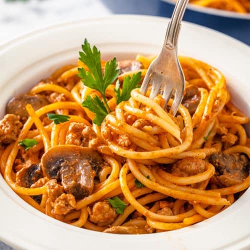 a white bowl with spaghetti and beef and mushrooms in it with a fork and parsley sprigs.