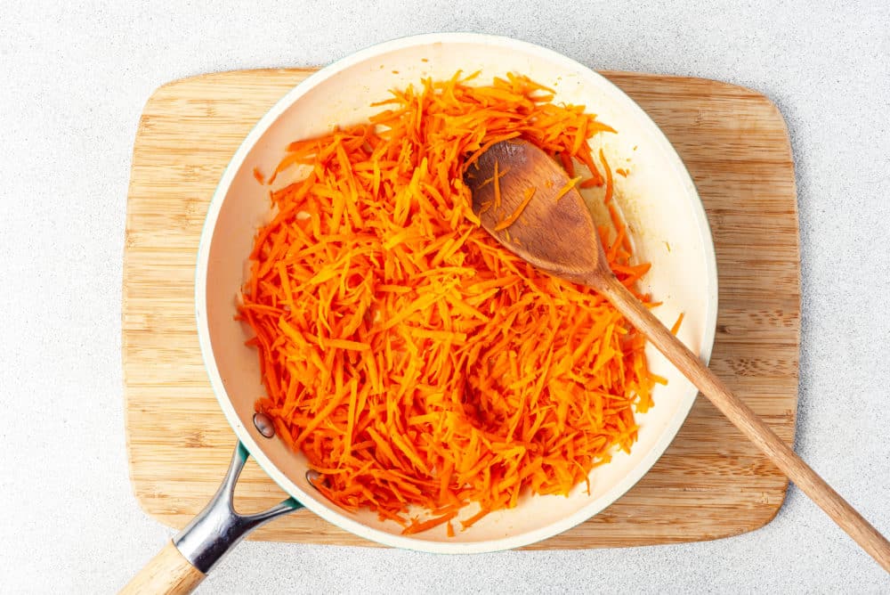 grated and sauteed carrot in a skillet with a wooden spoon on a wooden board.
