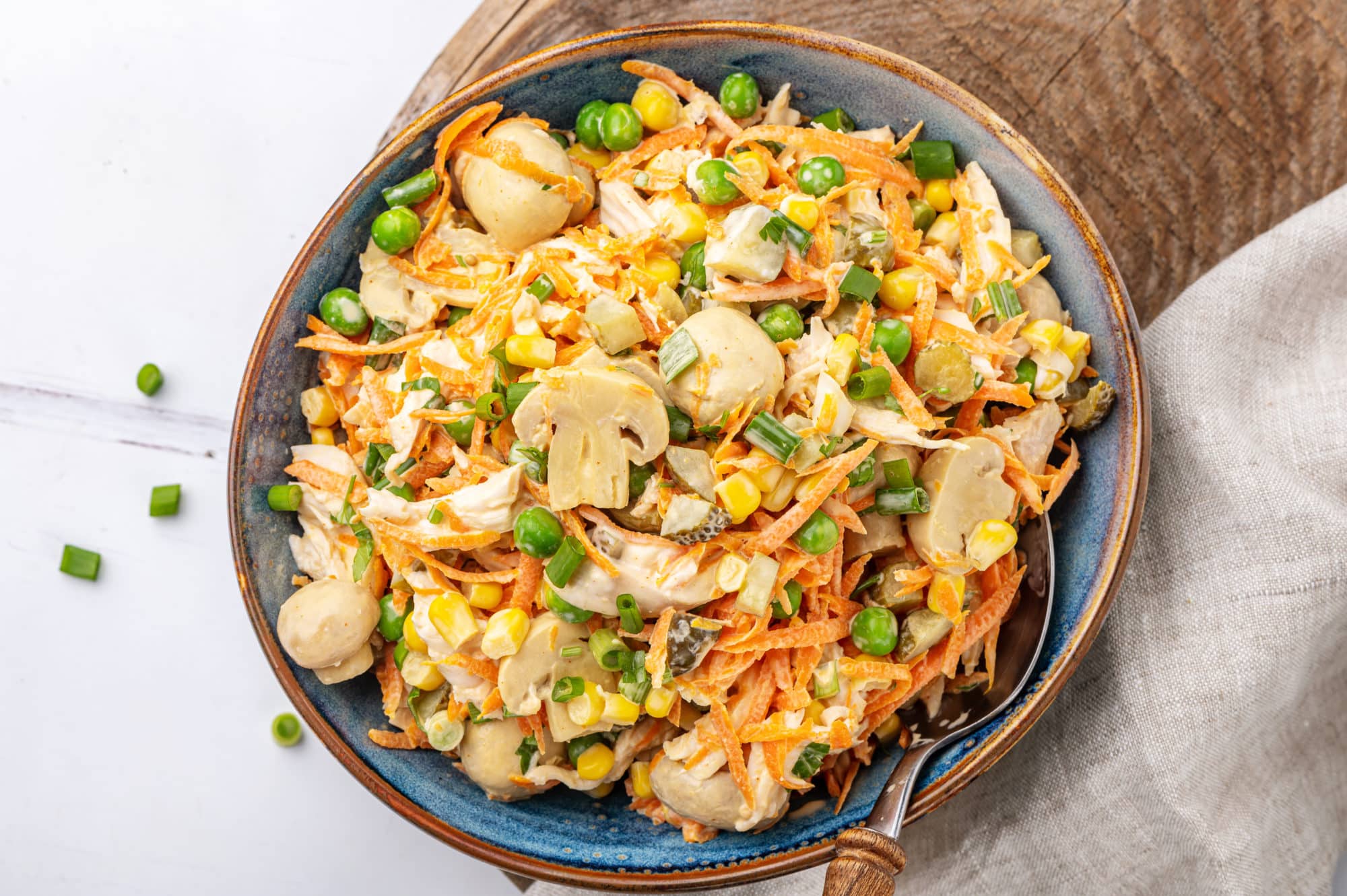 a spoon in a blue bowl filled with chicken veggie mushrooms salad standing on a brown board with a grey towel on the side.