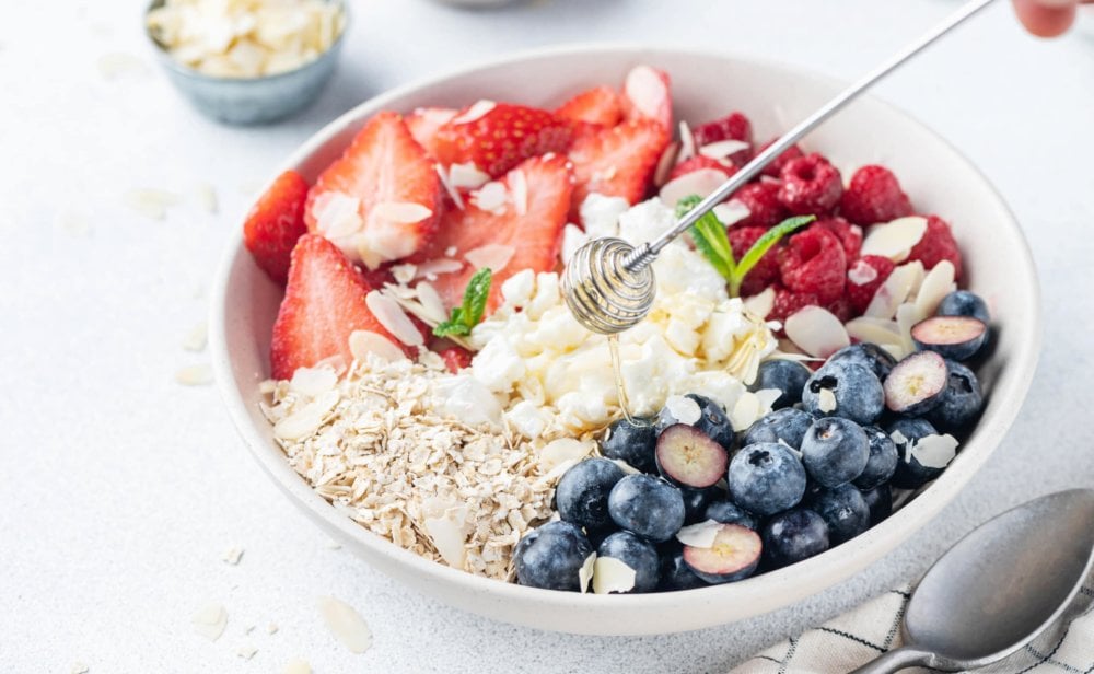 A bowl of cottage cheese and fruit with oats and silvered almonds and honey being drizzled on top.