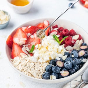 A bowl of cottage cheese and fruit with oats and silvered almonds and honey being drizzled on top.