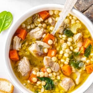 chicken soup with couscous and vegetables in a white soup bowl and silver spoon with bread on the side.