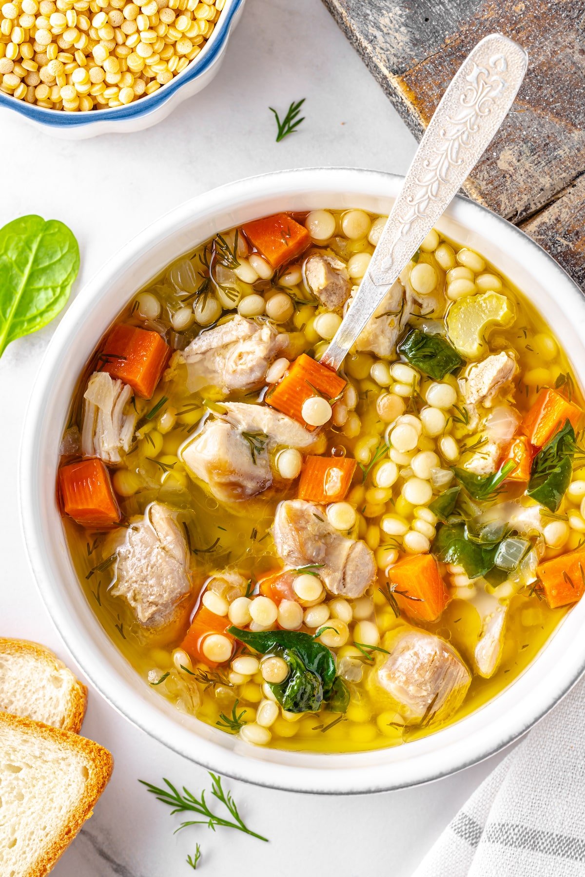 chicken soup with couscous and vegetables in a white soup bowl and silver spoon with bread on the side.