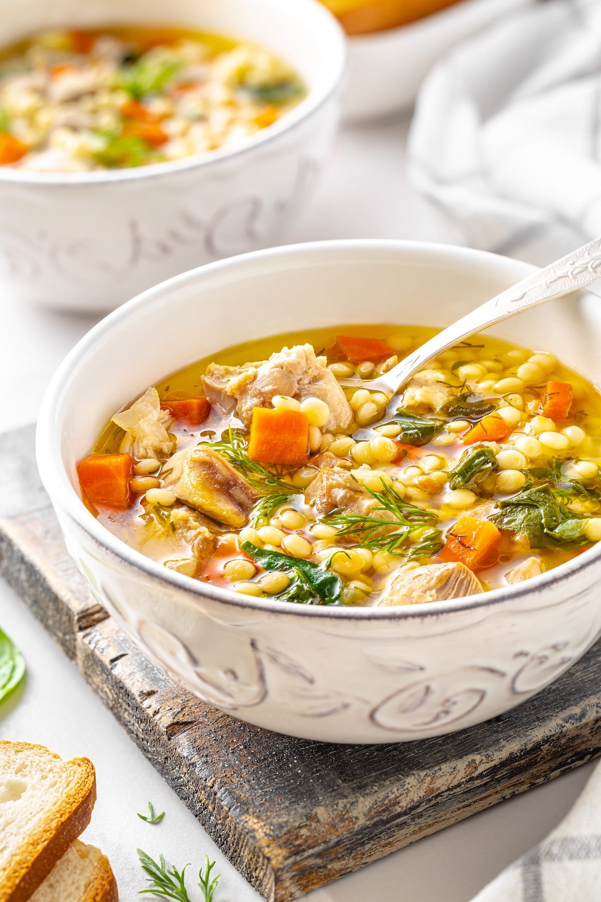 white bowl of chicken couscous soup with onion carrots fresh spinach and dill on a wooden board and bread on the side.