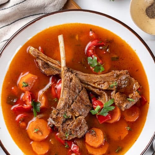 lamb chops in tomato water with herbs and chopped vegetables in a white bowl.