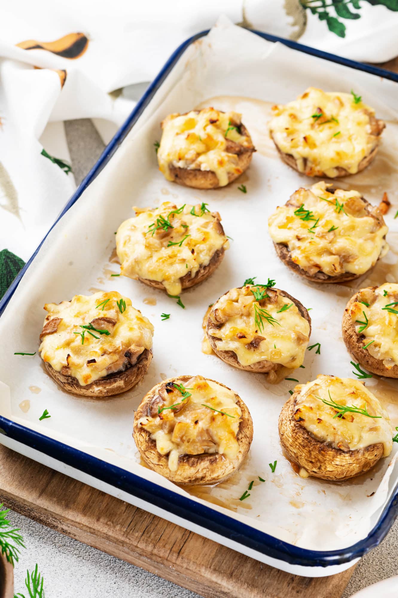 wooden-board-with-baking-dish-with-parchment-paper-and-stuffed-mushrooms-with-cheese-and-dill-and-a-white-towel