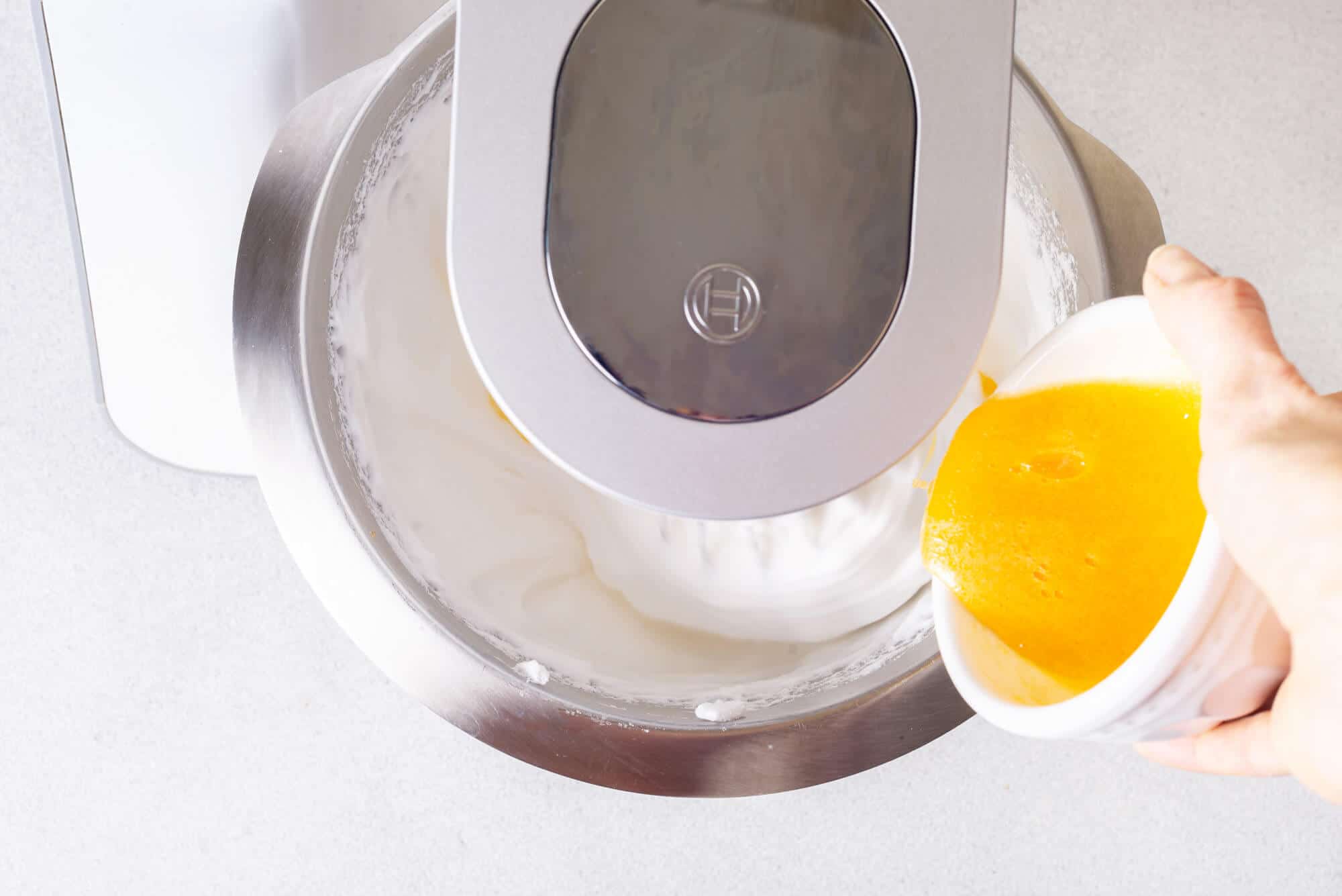 A stand mixer whisking egg whites and a hand pouring jello mixture into the mixer bowl. 