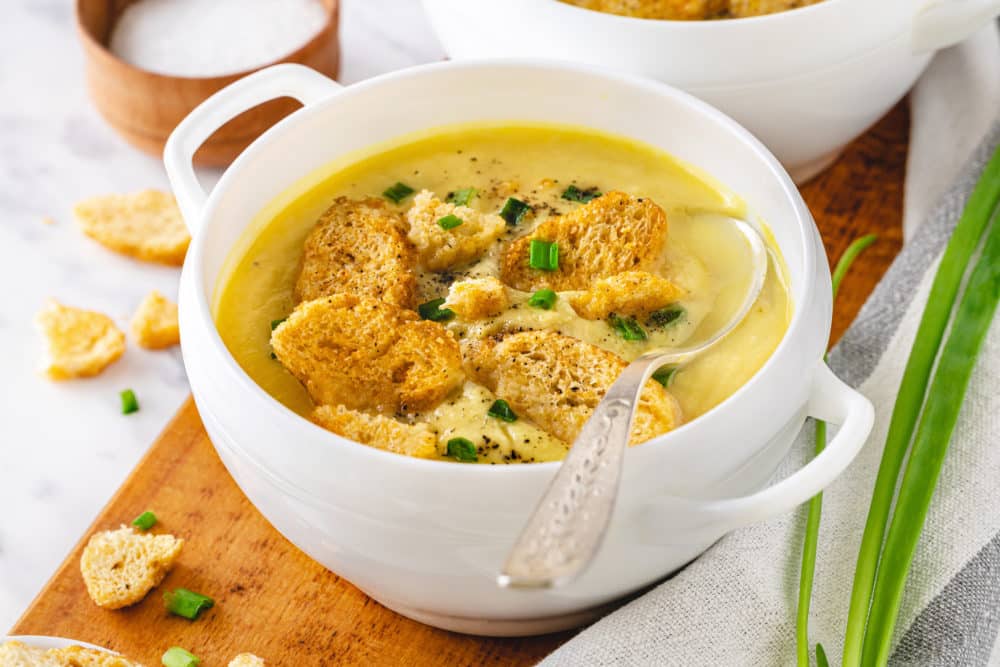 cauliflower-soup-in-a-white-bowl-with-croutons-on-top-and-with-a-spoon-on-a-wooden-board