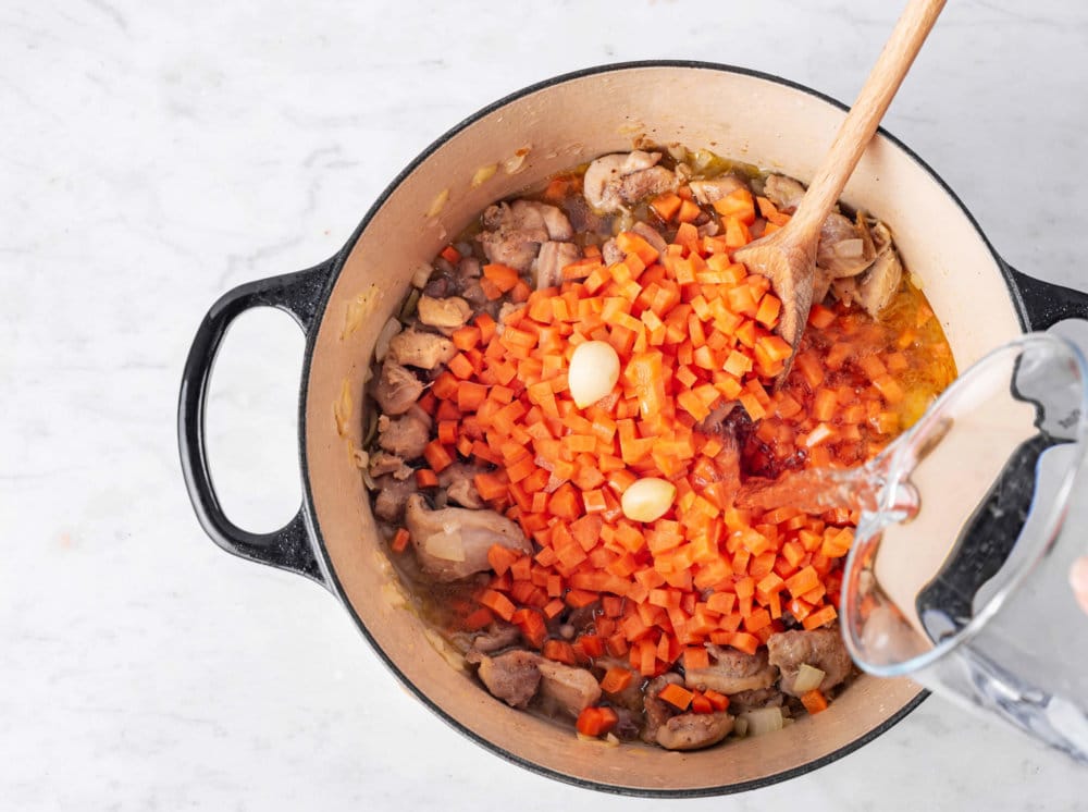 chopped-carrots-chicken-garlic-and-water-being-added-to-a-pot-with-a-wooden-spoon