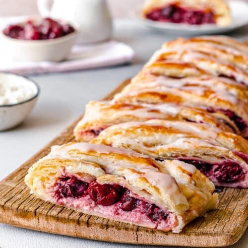 cherry cheese danish with glaze on top on a wooden board cut up into slices.