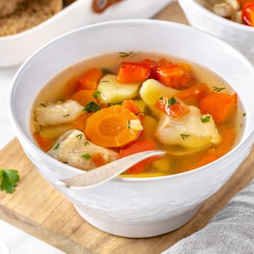 a white bowl of fish soup with veggies floating in it on a wooden board with a spoon in the soup.