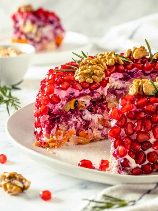 layeredpomegranate salad wreath shaped with a slice missing on a white plate with rosemary on top and around.