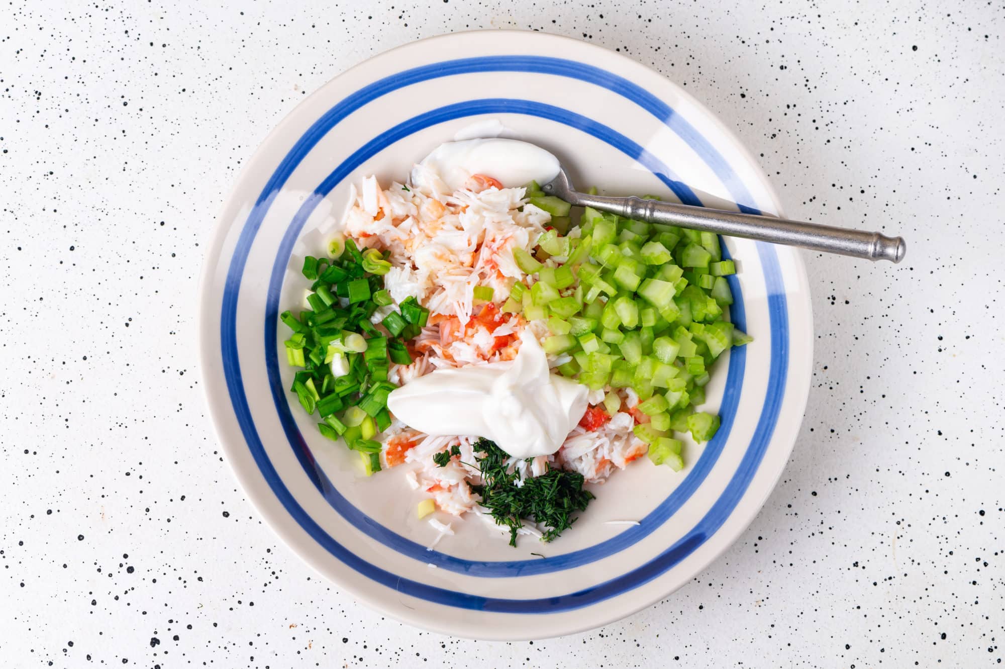 ingredients of a crab salad snack in a white bowl with blue stripes with a spoon.