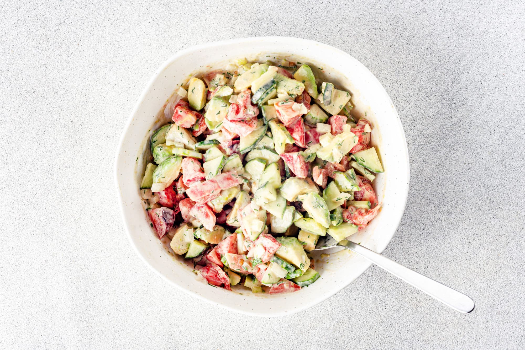 prepared avocado tomato salad with cucumbers in a white bowl with a spoon.