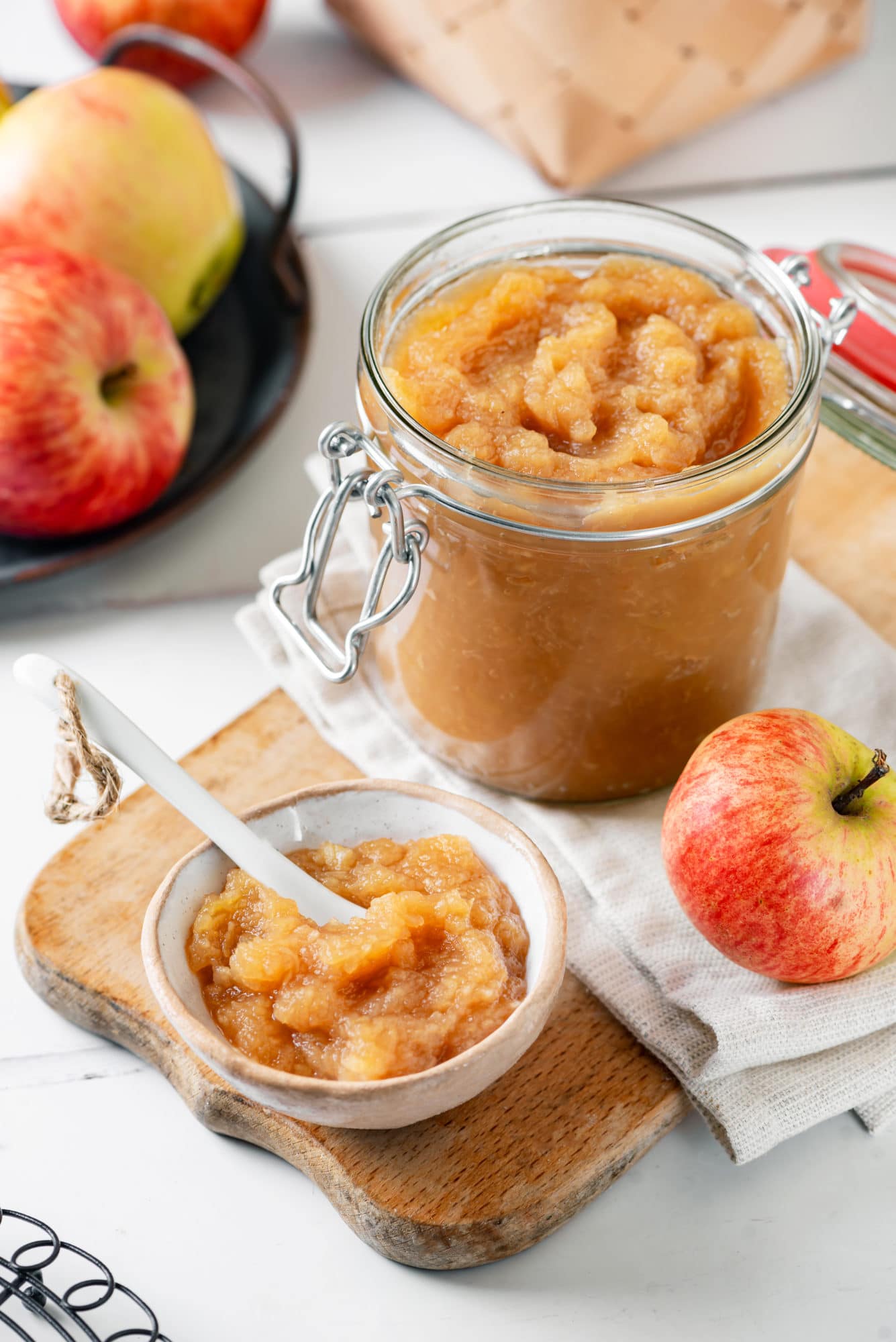 apple-jam-in-a-clear-jar-and-a-small-bowl-with-a-spoon-on-a-wooden-board-with-apples-on-the-side