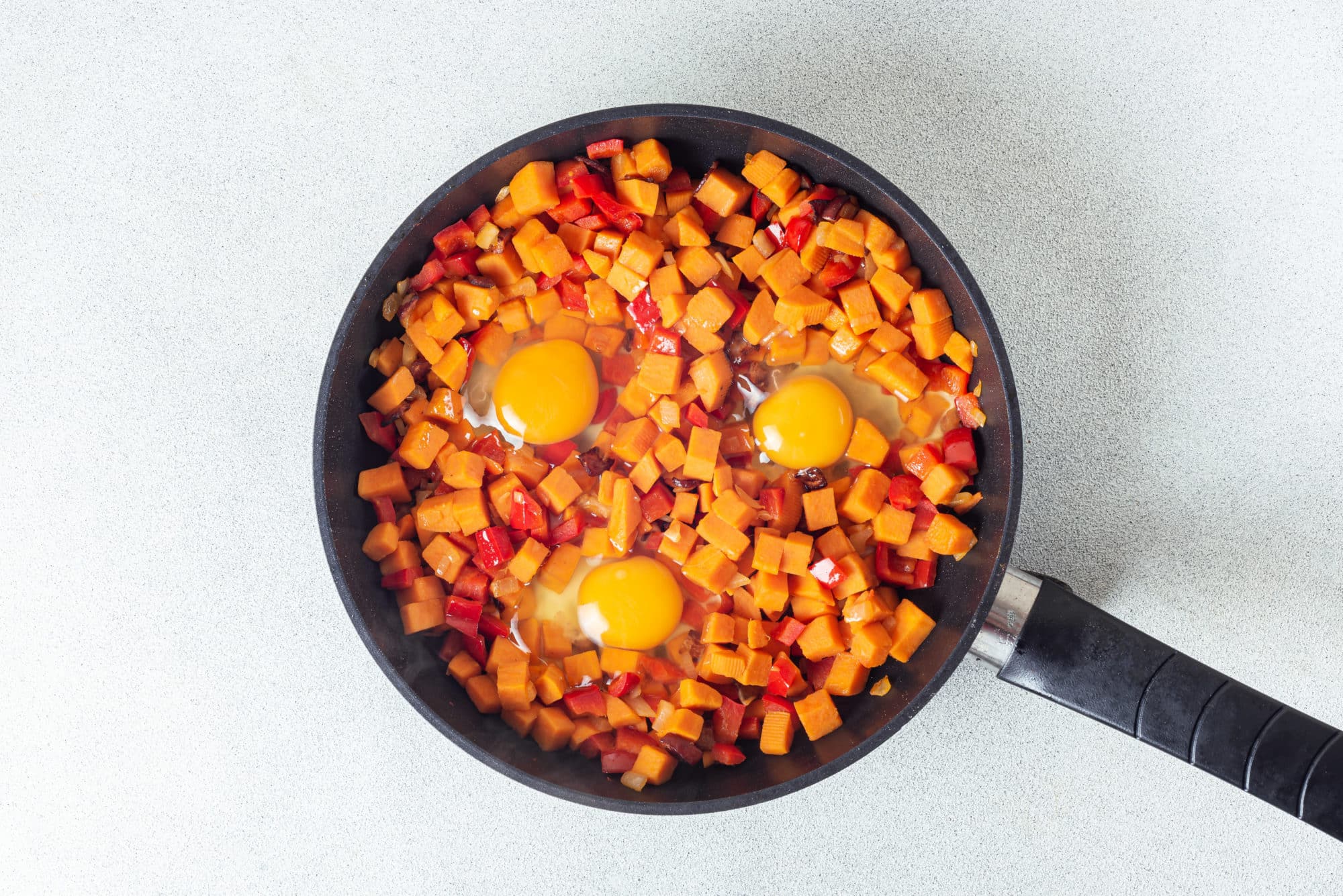 sweet potatoes and red bell peppers with cracked raw eggs in a black pan.