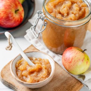 A glass jar and a white bowl of apple jam with a spoon in the bowl and apples all around.