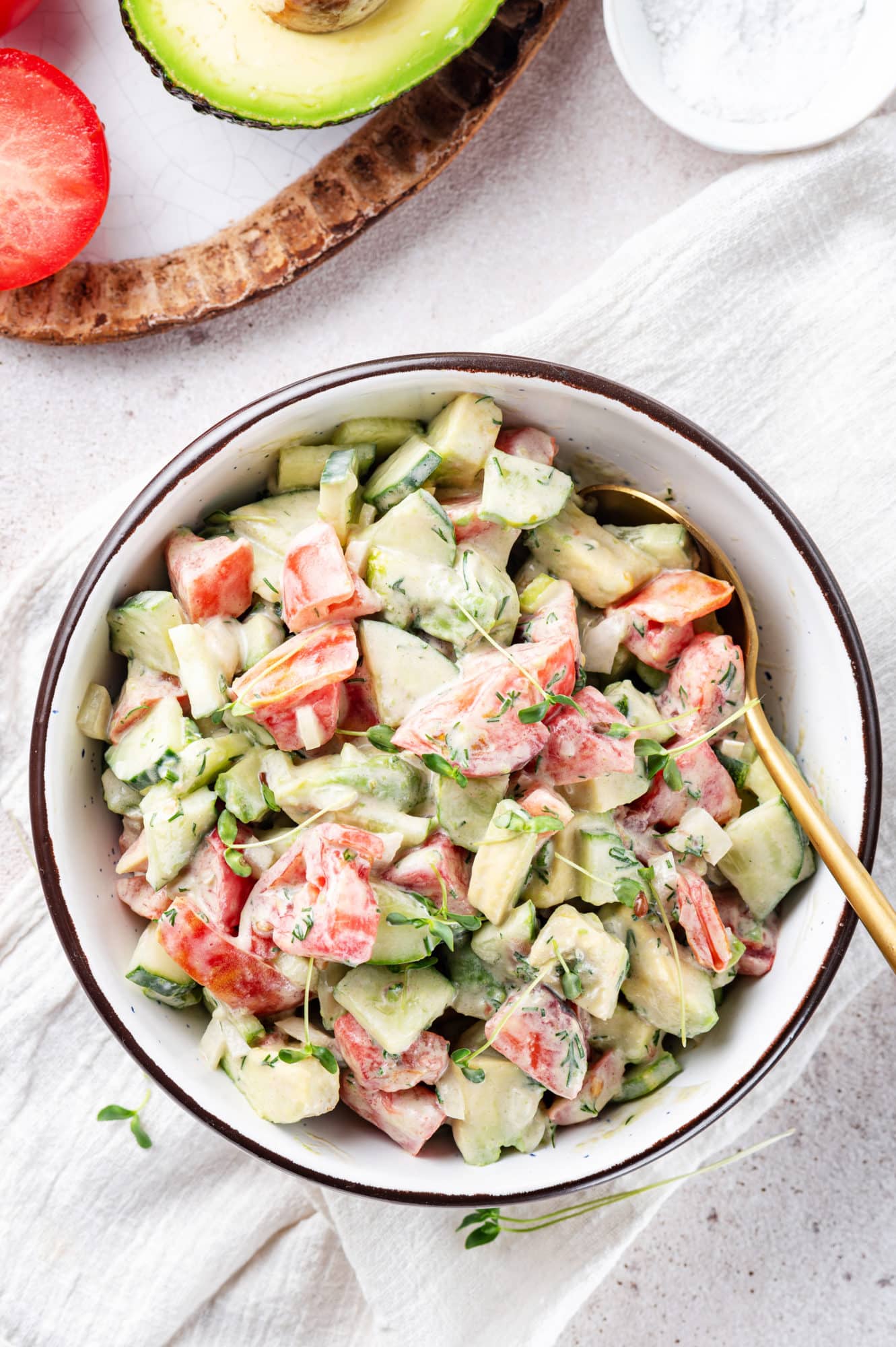 a gold spoon in a white bowl with avocado tomato and cucumber salad in it.