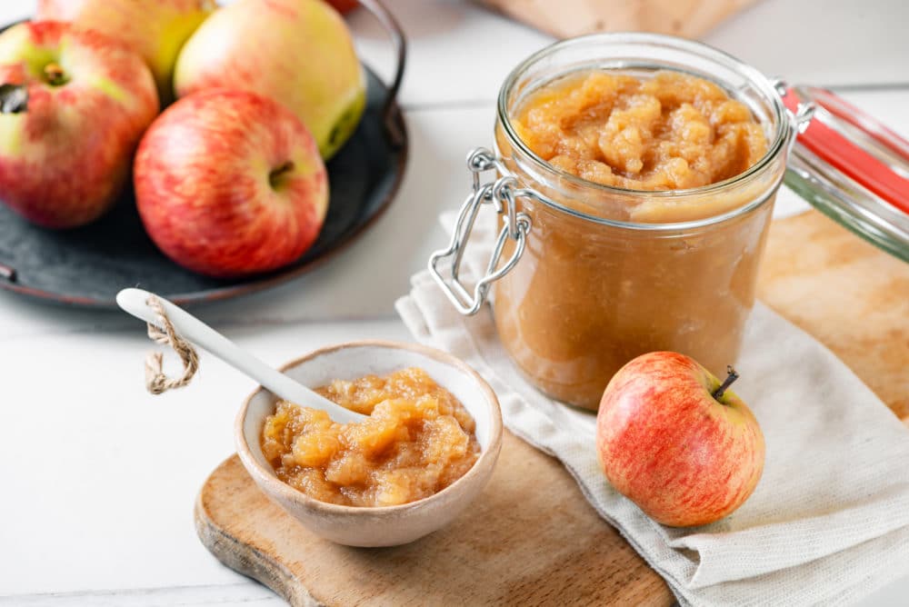 apple jam in a glass jar with a small bowl of jam, a spoon, and an apple on the side.