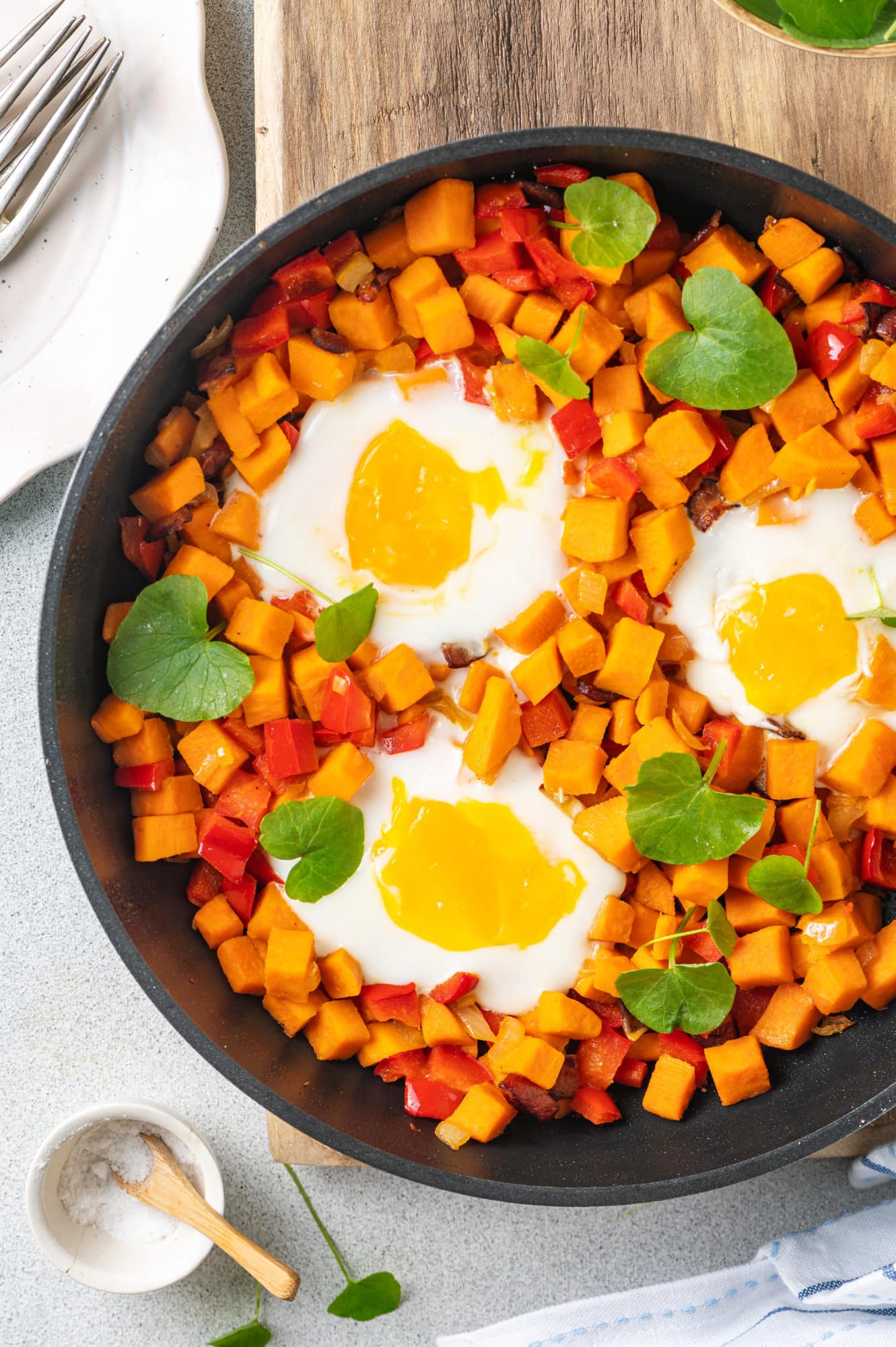 wooden-cutting-board-under-a-black-skillet-filled-with-red-pepper-sweet-potatoes-runny-eggs-and-watercress-and-silver-forks-on-a-white-plate-and-a-small-bowl-of-salt-with-a-small-wooden-spoon-and-a-striped-towel
