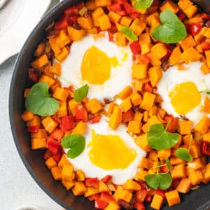 Sweet potato and eggs in a skillet with a sprinkle of micro greens on a wooden board and forks and a small bowl of salt on the side.