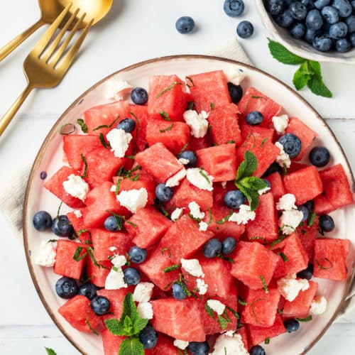 Watermelon feta salad on a white plate topped with fresh mint and a fork, spoon, and a bowl of blueberries on the side.