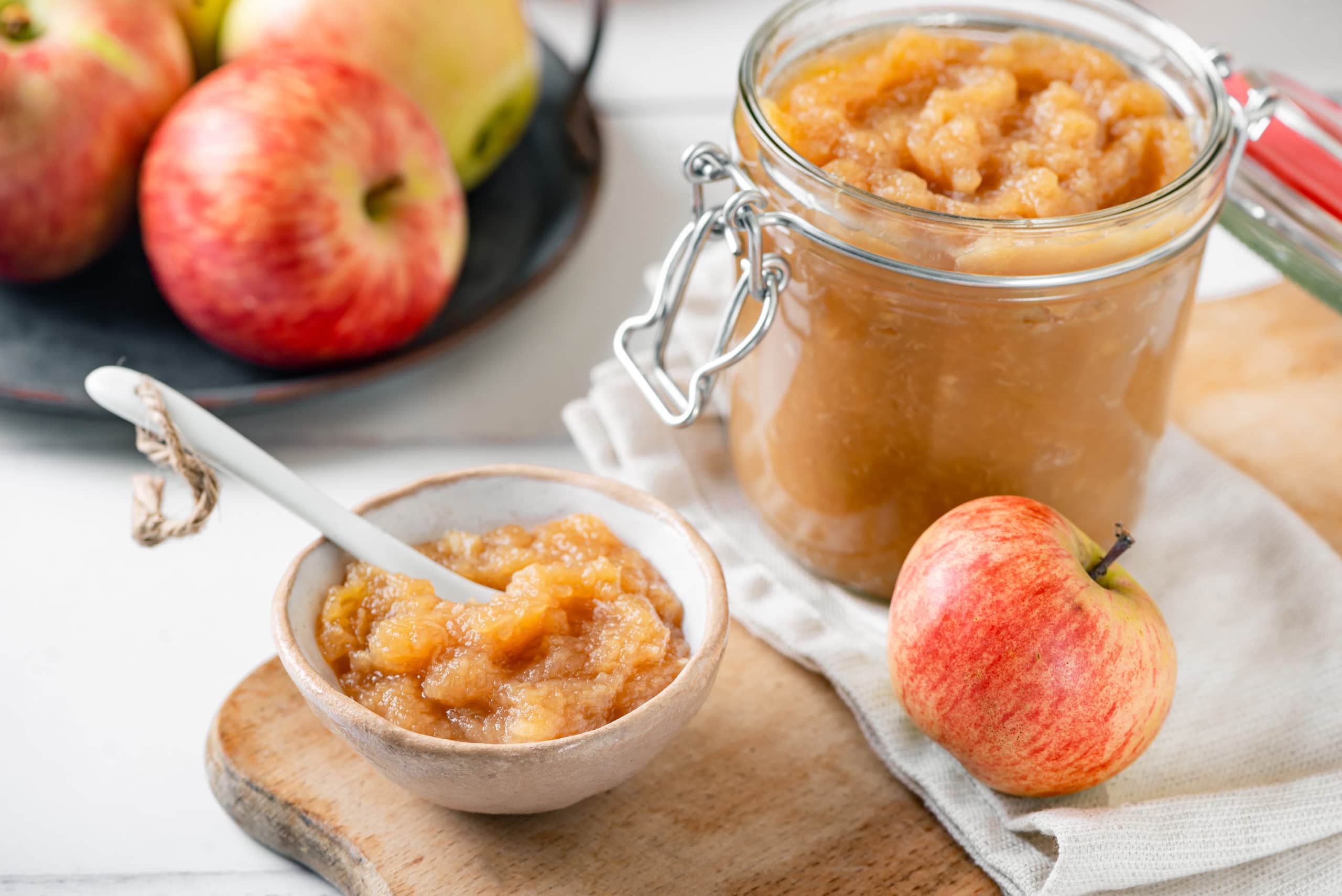 A glass jar and a white bowl of apple jam with a spoon in the bowl and apples all around.