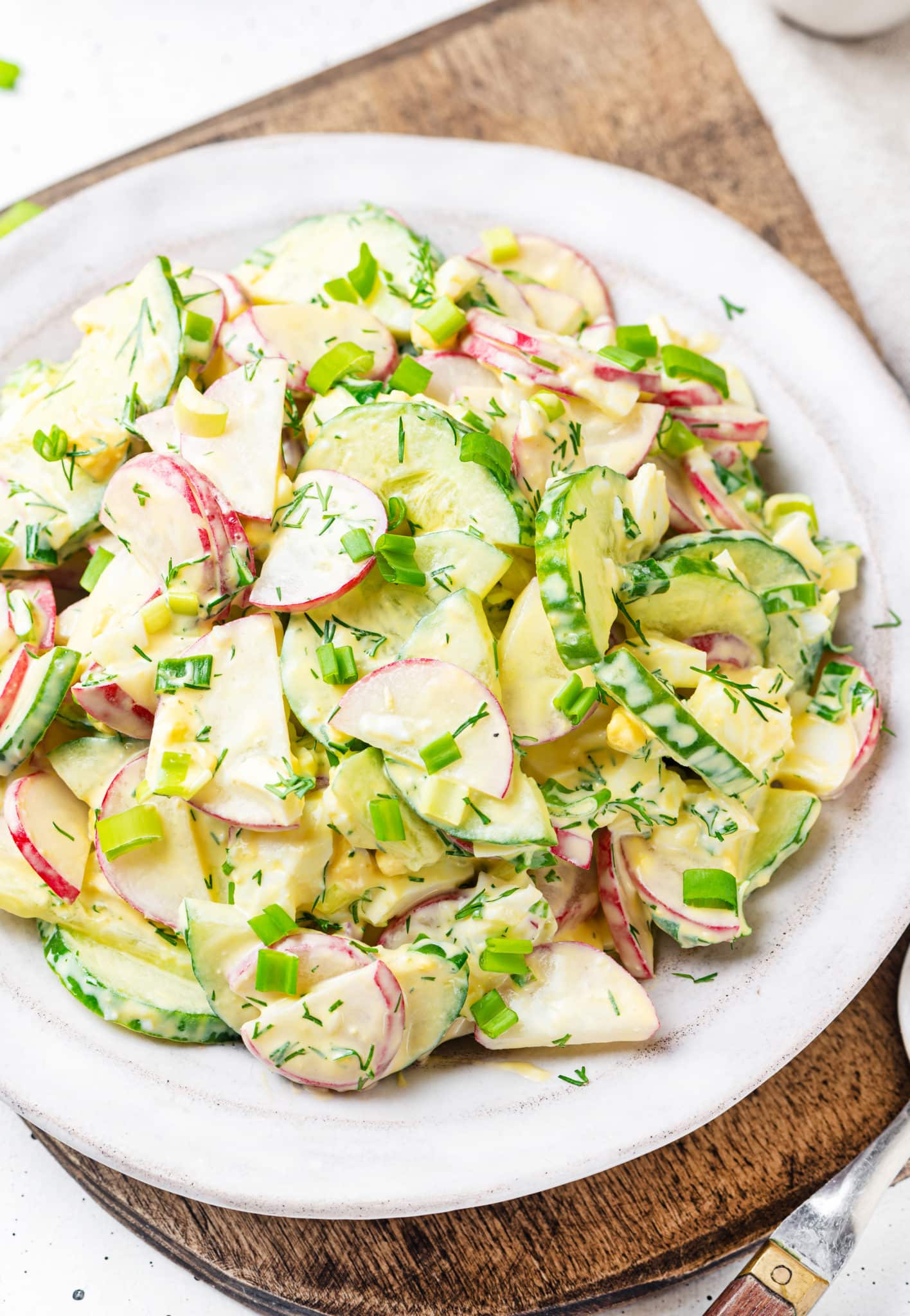 wooden-board-under-white-plate-with-radish-cucumber-salad-with-dill-and-green-onion-and-dressing-and-a-silver-spoon