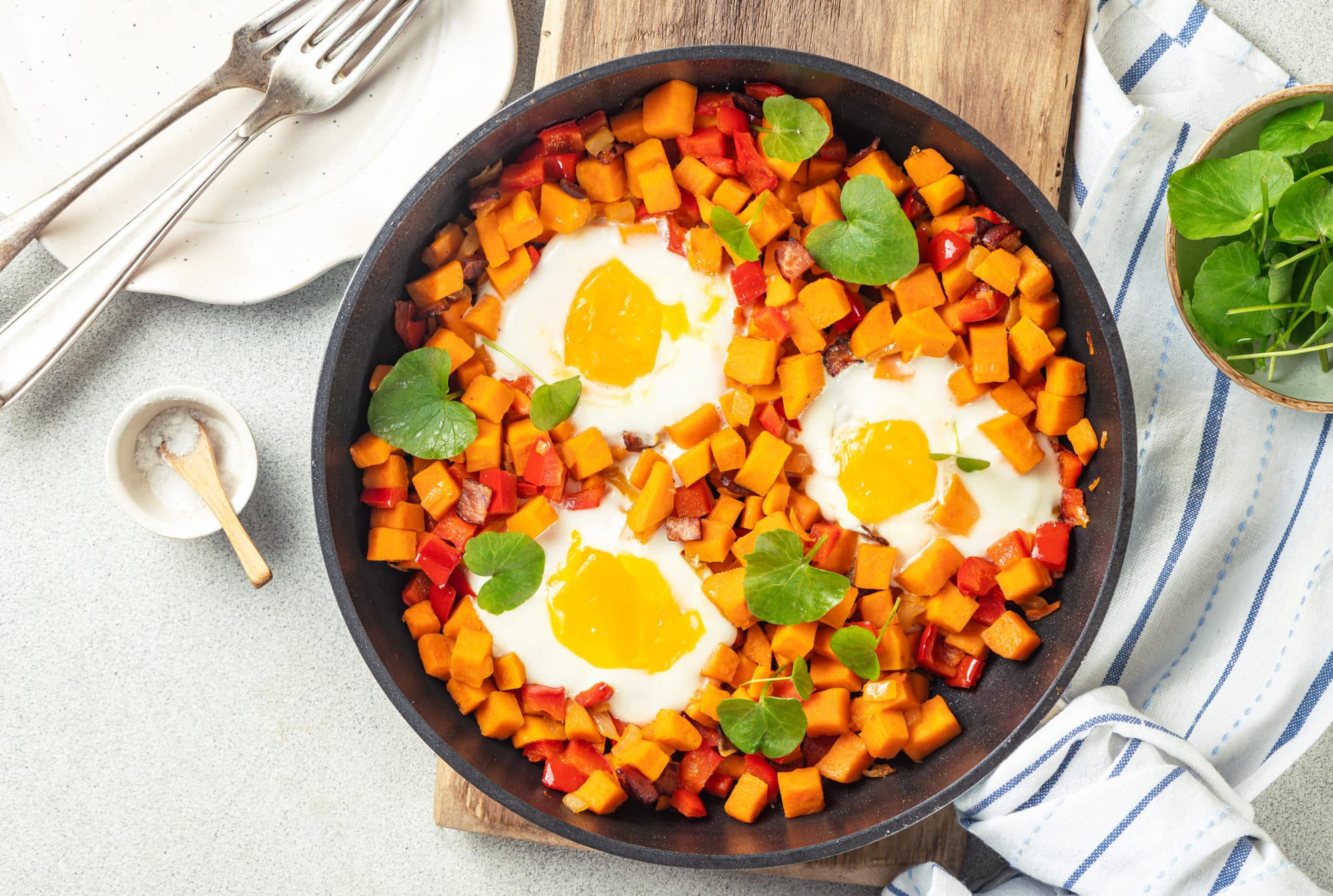 wooden-cutting-board-under-a-black-skillet-filled-with-red-pepper-sweet-potatoes-runny-eggs-and-watercress-and-silver-forks-on-a-white-plate-and-a-small-bowl-of-salt-with-a-small-wooden-spoon-and-blue-and-white-striped-towel-under-a-white-bowl-filled-with-watercress
