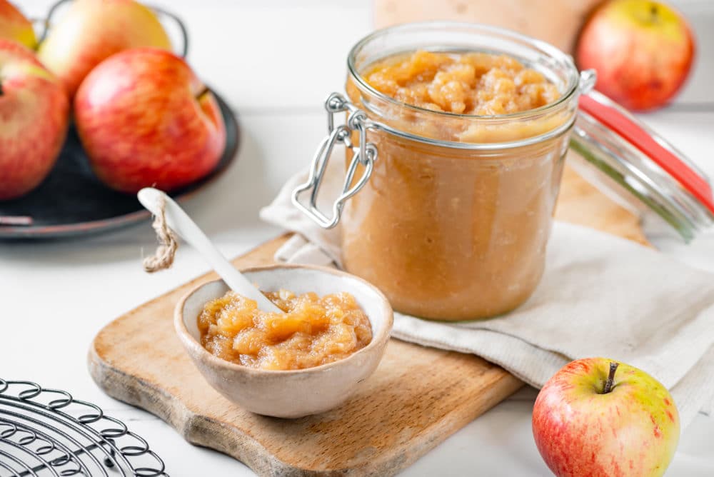 apple jam in a jar and some in a bowl on a board with a spoon.