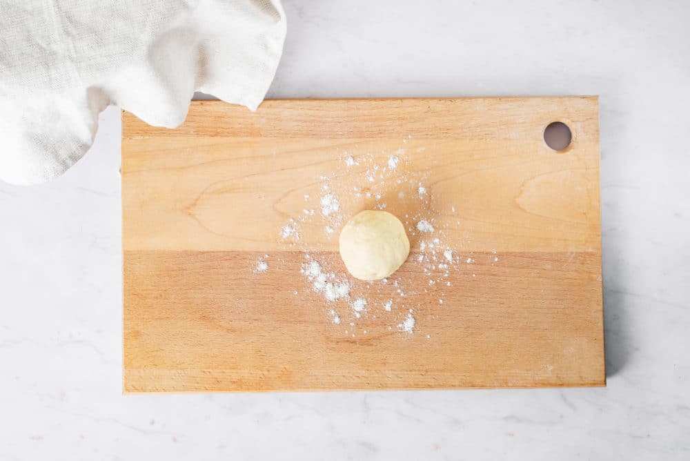 A little ball of dough on a wooden cutting board with some flour sprinkled around.