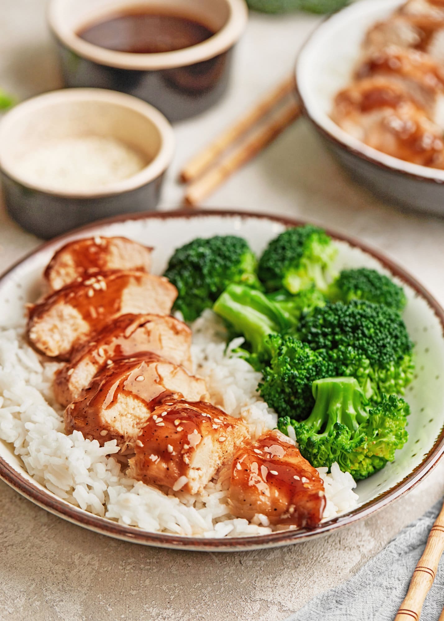 A bowl of sliced teriyaki chicken with broccoli and rice, and chopsticks on the side.