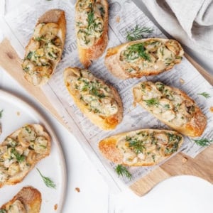 mushroom-crostini-on-wooden-cutting-board-with-paper-and-crostinis-on-white-plate