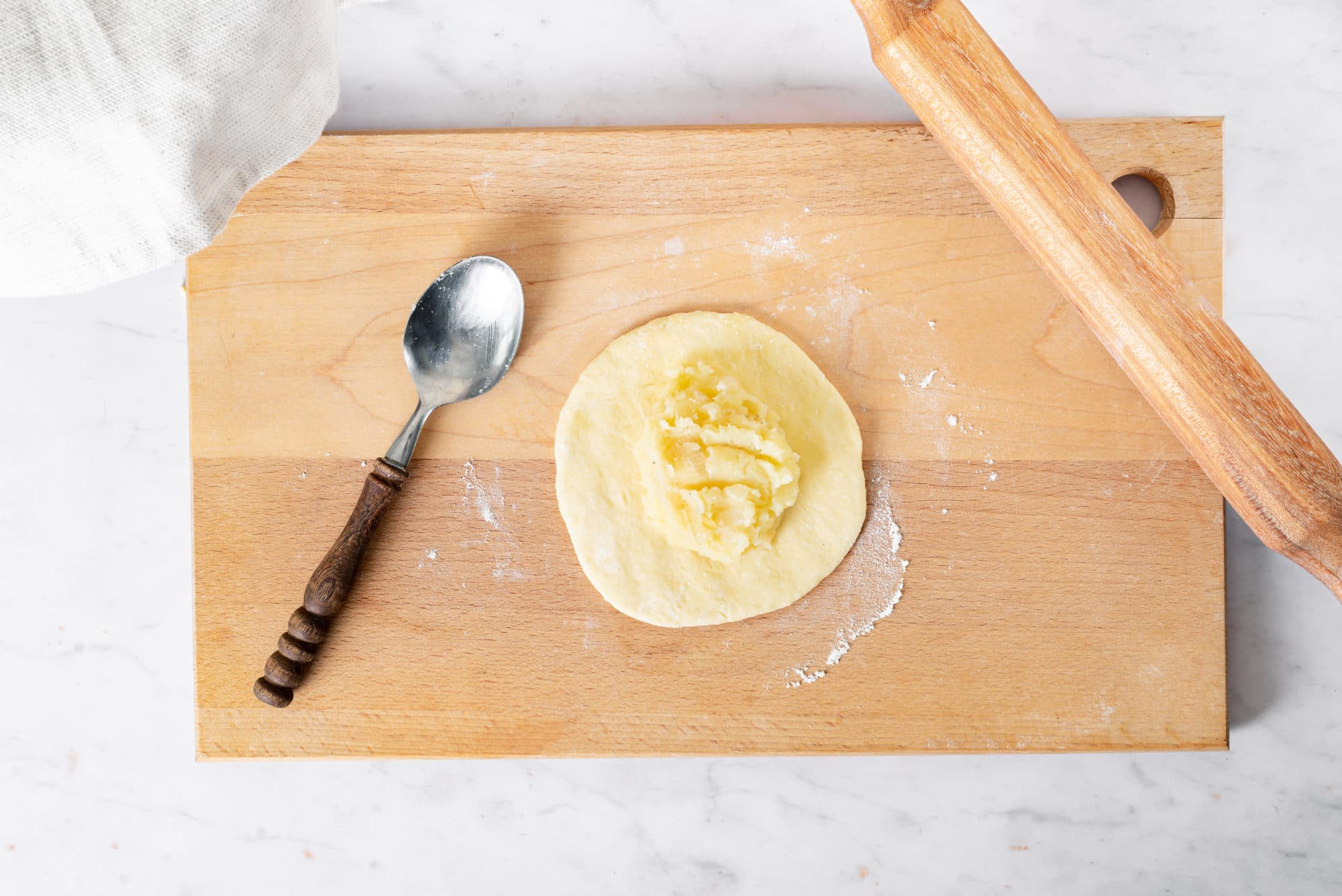 Rolled out dough on a wooden cutting board filled with mashed potato filling with a wooden rolling pin and a spoon on the side.
