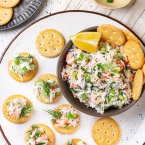 snack of crab salad in a brown bowl and more crackers on a white plate with small amounts of crab salad on the crackers.