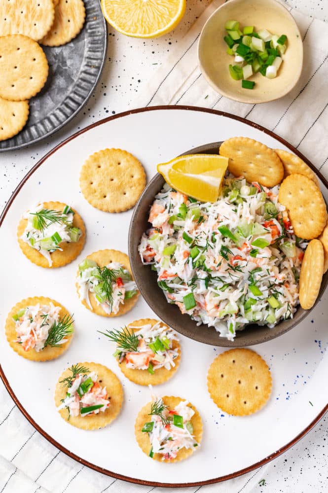 snack of crab salad in a brown bowl and more crackers on a white plate with small amounts of crab salad on the crackers.