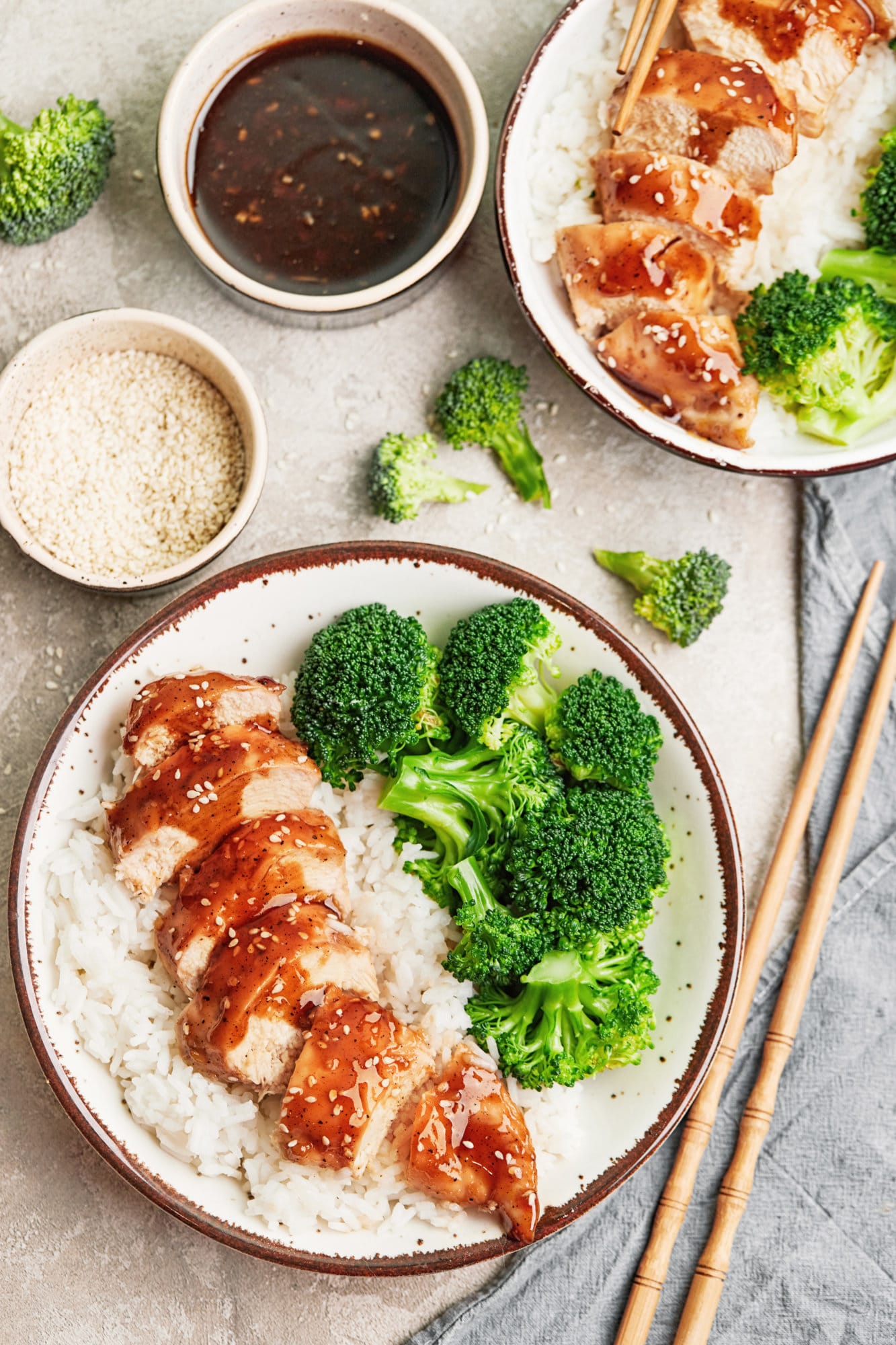 Two bowls of sliced teriyaki chicken over a bed of rice with broccoli and chopsticks on the side.