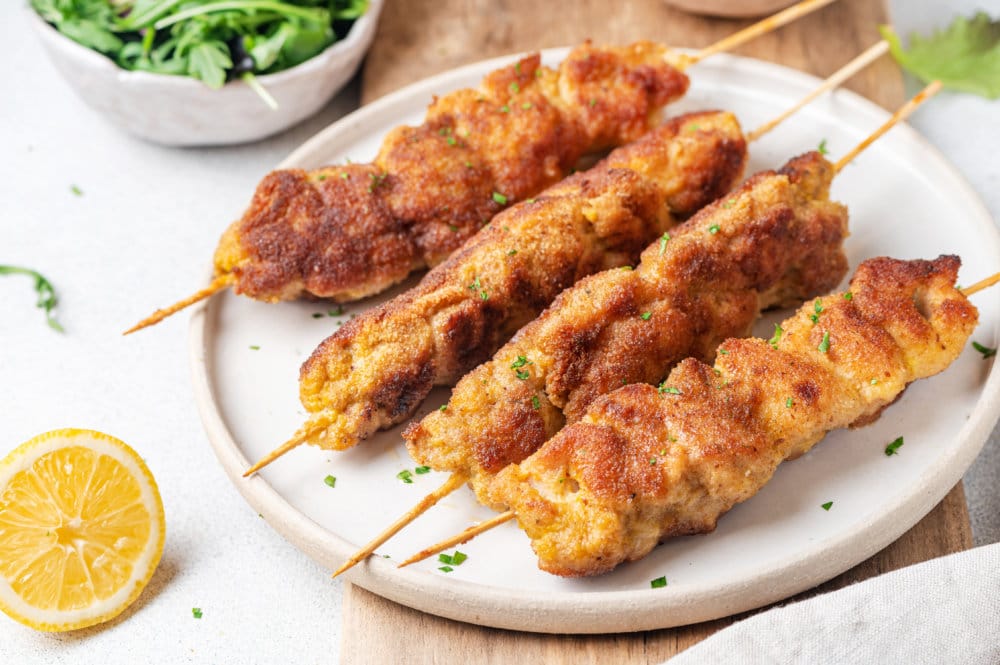 white-plate-with-four-chicken-skewers-on-a-brown-board-lemon-wedge