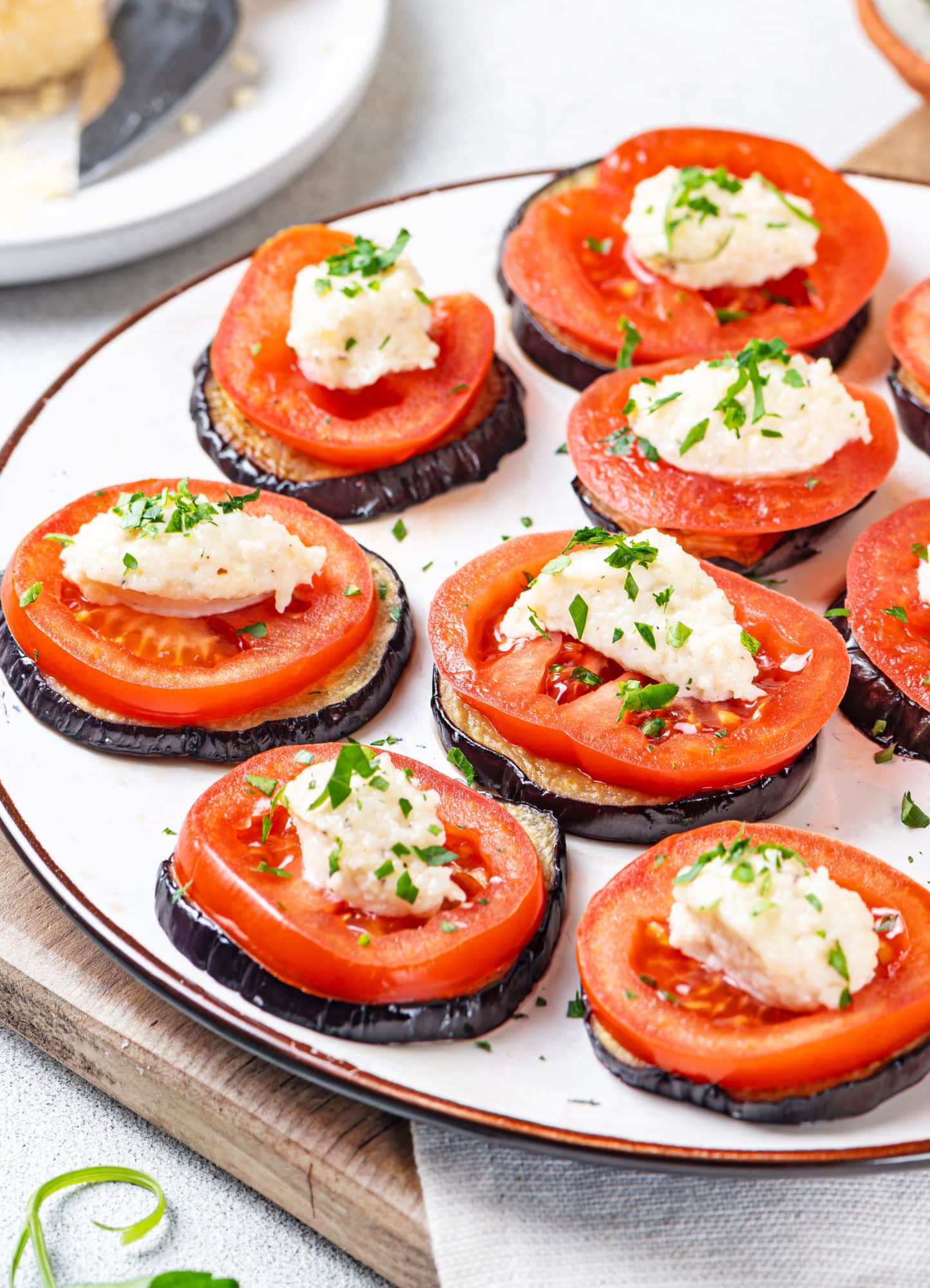 eggplant and tomato stacked appetizer laid out slices on a white plate on a brown cutting board and a grey towel.