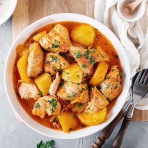 braised-chicken-potato-dish-in-a-white-bowl-on-a-wooden-board-with-a-fork-and-a-spoon-and-a-white-towel