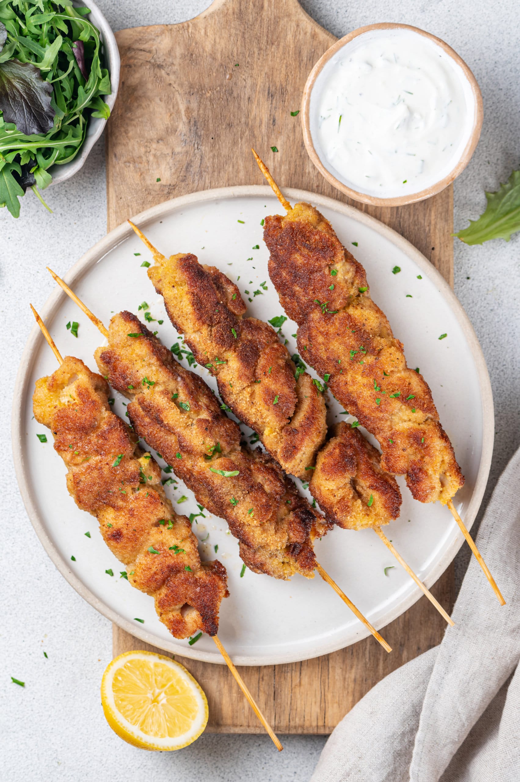 A white plate witt four chicken skewers on a brown board with a towel and lemon on the side. 