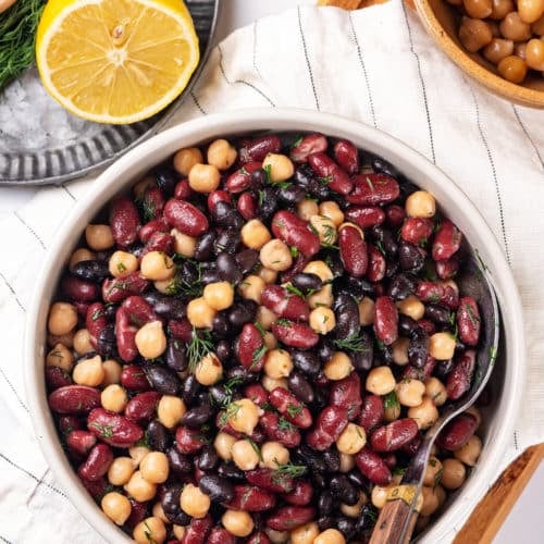 a mixed bean salad in a bowl on a white towel with a spoon in the bowl and a lemon wedge and a brown bowl of chickpeas on the side.