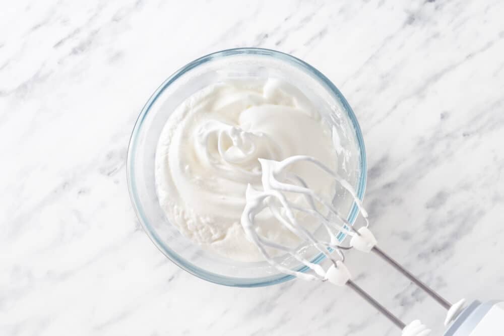 Beaten egg whites in a glass bowl with mixers.