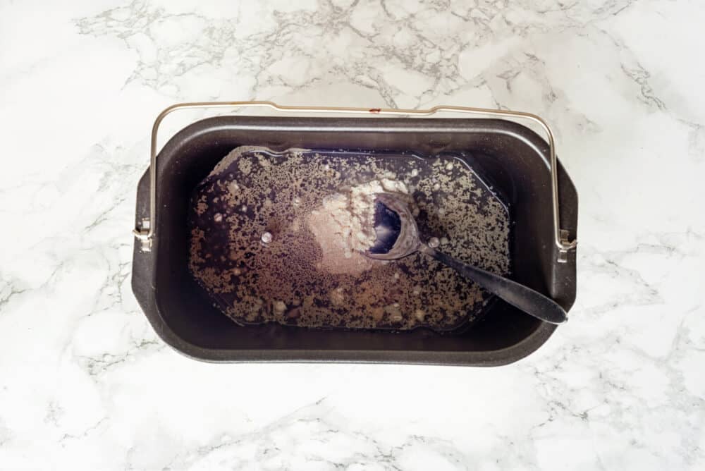 Bread ingredients in a bread machine bucket mixing with a spoon.