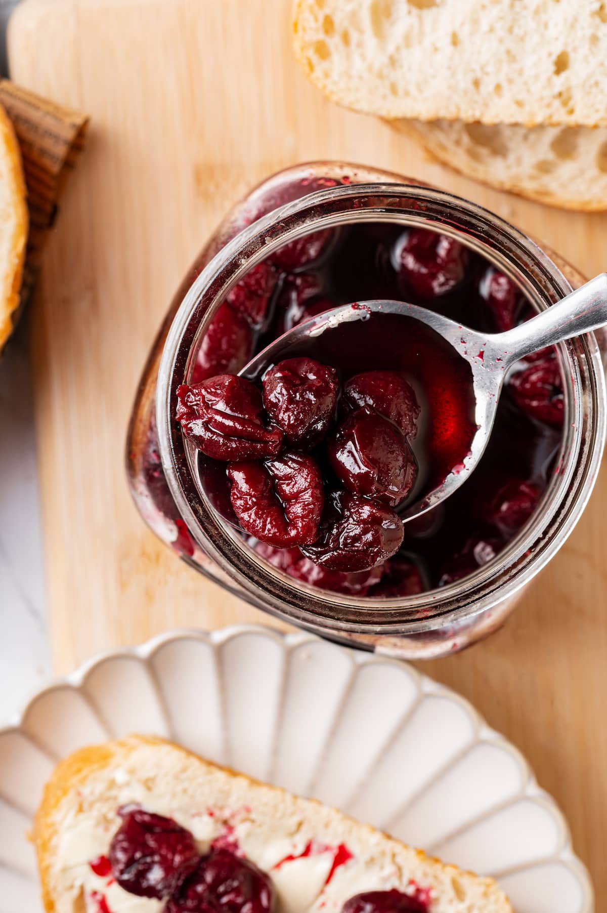 cherry jam in glass container with silver spoon on wooden board, with a slice of bread on the side on a white plate with butter and cherry jam.