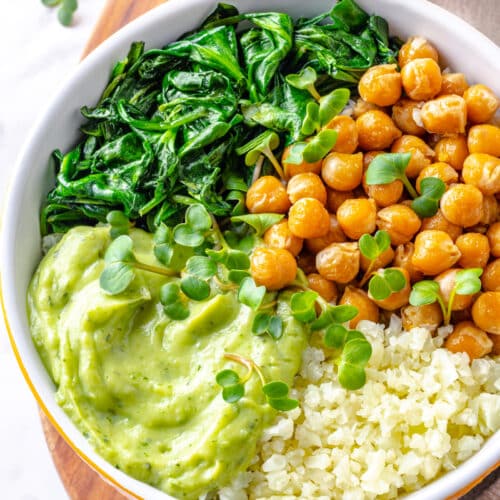 chickpea buddha dish in a white and brown bowl on a board with a towel and guacamole on the side.