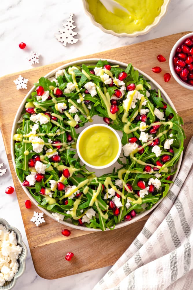 green salad in a wreath shape on a white plate with a small white bowl of avocado dressing in the center all on a brown board with pomegranate seeds and snowflake decorations sprinkled around and a striped white grey and brown towel on the side.