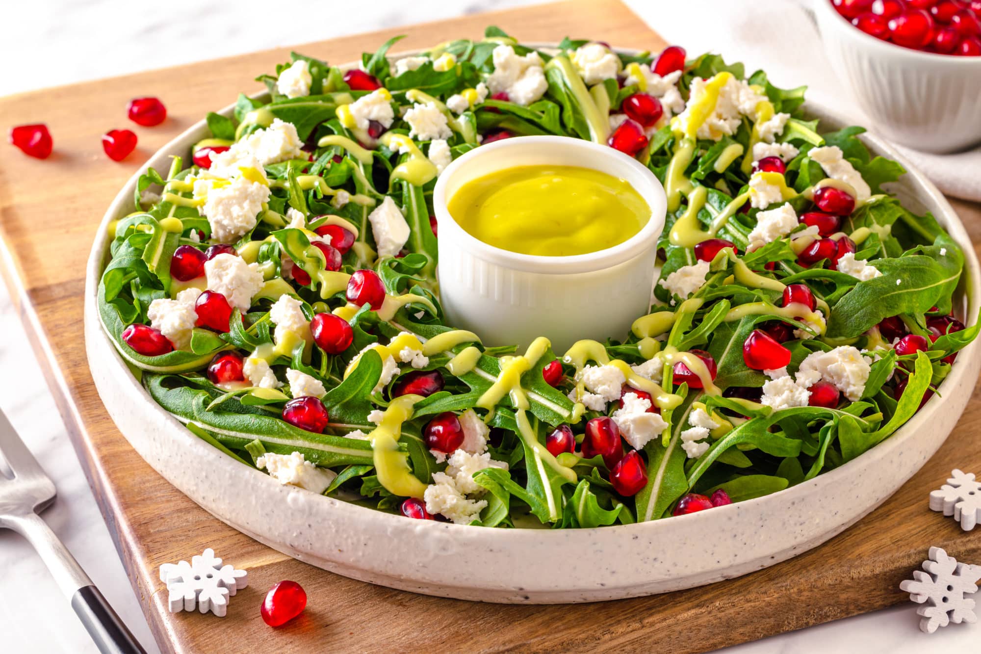 a white plate with a green salad and drizzled green dressing on top and dressing in a small white dish in the center with pomegranate seeds and feta cheese sprinkled on top of the salad.